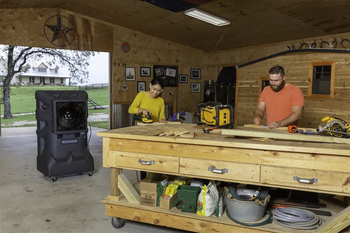 Homeowners working at a large wooden mobile workbench in a well-lit garage workshop with a portable cooler and power tools